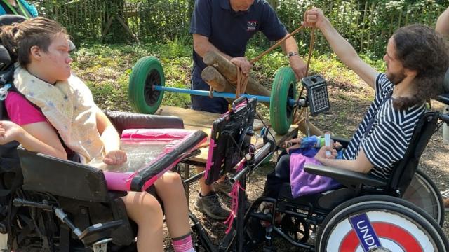 Two Treloar's students outdoors in the woods engaging with a man demonstrating a green-wheeled device in a wooded area. The students are looking carefully.