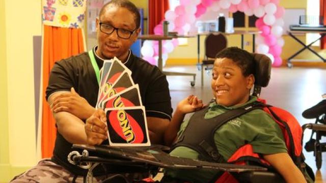 Treloar's student with his assistant playing UNO card game in a brightly decorated room with pink balloon arch in the background. The assistant is holding a big size cards and the student is looking at them with a smile.