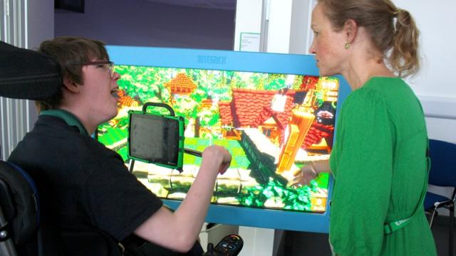 Treloar's student in front of a big screen displaying a colourful video game with a teacher in a green dress having a discussion about the game.