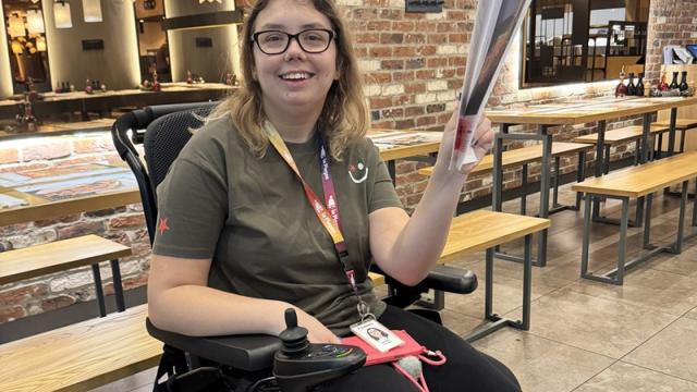 Treloar's student in an empty restaurant holding a big menu and smiling to the camera. She is wearing glasses and a work experience uniform.