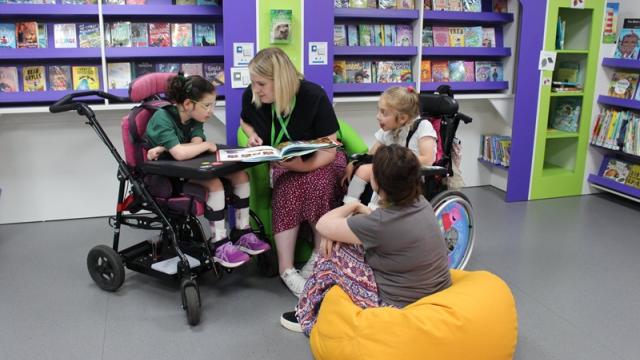 Teacher reading a book to two Treloar's primary students and a support assistant sitting on a yellow beanbag in a colourful library.