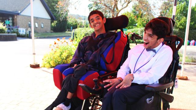 Two Treloar College student smiling and enjoying a sunny day outdoors near a building and garden. They are wearing a formal attire. 