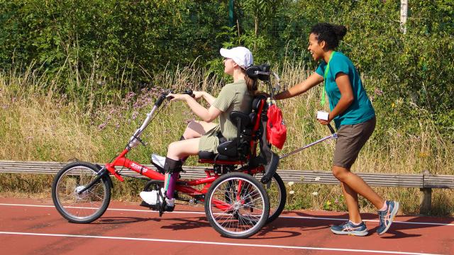 A physiotherapist pushes Treloar's student in an adapted tricycle along a track surrounded by greenery. It is a hot day, both women are wearing shorts and T-shits.