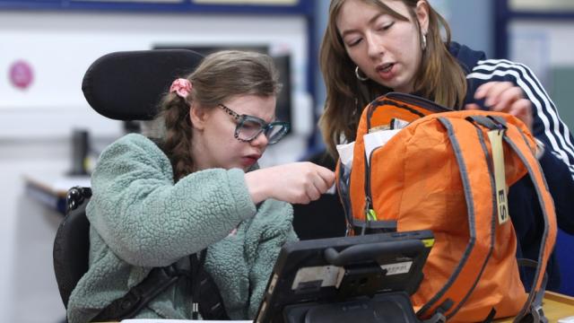 Treloar College student is trying to pack her bright orange backpack with assistance from her assistant; there is a tablet in front of her on the desk. She is very focused on the task.