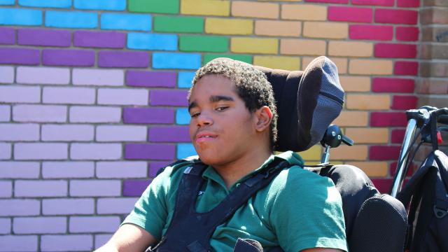 Treloar School student wearing a green T-shirt uniform and poses in front of a colourful brick wall painted in purple, blue, yellow and red hues.