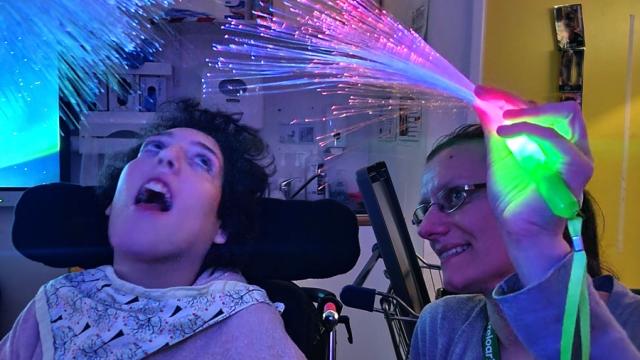 Treloar College student enjoying colourful fiber optic lights held by a smiling assistant in a sensory room. Lights are dimmed and the student is mesmerised by the fiber lights.