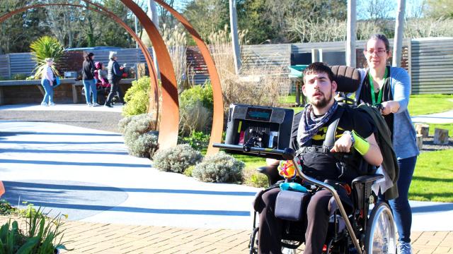 Treloar College student in a garden, being pushed by his assistant along a sunny outdoor path with modern sculptures and greenery in the background. His AAC device is attached to his wheelchair.