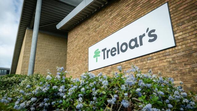White rectangular sign with 'Treloar's' and a green tree logo mounted on a brick building wall above flowering bushes.