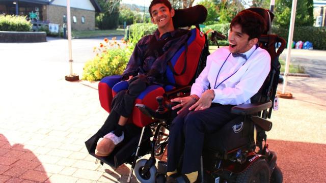 Two Treloar College student smiling and enjoying a sunny day outdoors near a building and garden. They are wearing a formal attire. 
