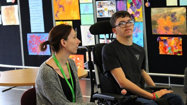 Treloar College student and his assistant with a green lanyard sit together in a room decorated with colourful abstract artwork on the walls. They are both looking serious.
