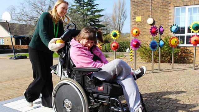 A student support assistant pushes a smiling Treloar College student outdoors near colourful flower-shaped musical instruments on a sunny day.