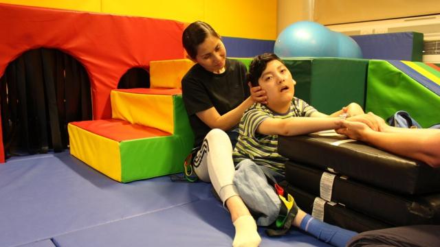 An assistant gently supports Treloar School student who is out of his wheelchair seated on padded mats in a colourful therapy room. The assistant is supporting his head by holding his head.