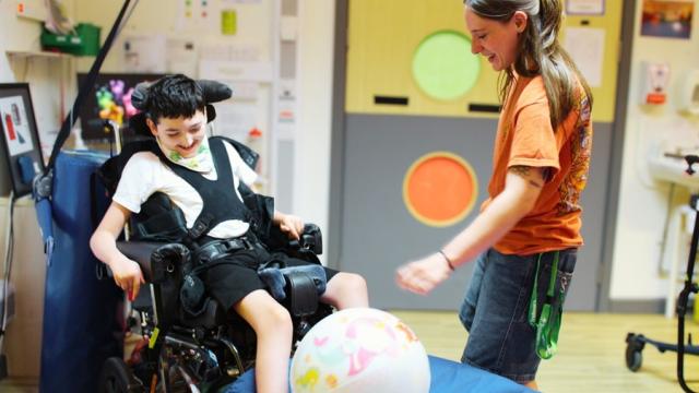 Treloar School student smiling as his assistant helps him kick a colourful ball indoors during a playful physiotherapy session. They are both having fun and are engaged in the activity.