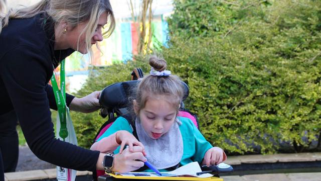 A student support assistant assists Treloar's primary student as she writes on a clipboard placed on the tray attached to her wheelchair. They are outdoors near greenery.