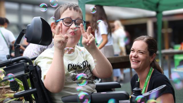Treloar's primary student making peace signs while surrounded by floating bubbles at an outdoor event with his assistant next to him, smiling.