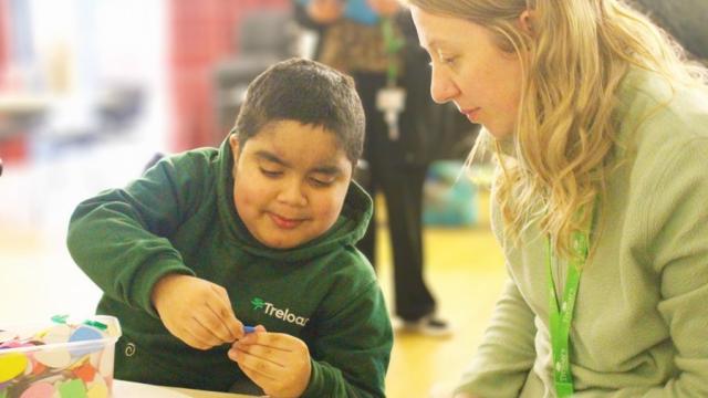 Treloar's primary student wearing a green hoodie uniform focused on a craft activity while his assistant with blonde hair attentively watches him at a table.