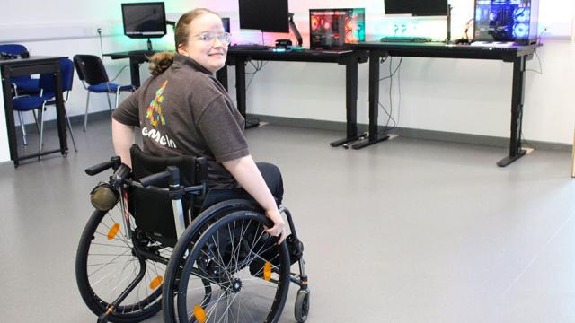 Treloar College student looking through her shoulder and smiling at the camera. She is in a computer room with multiple desktop setups on a long table.