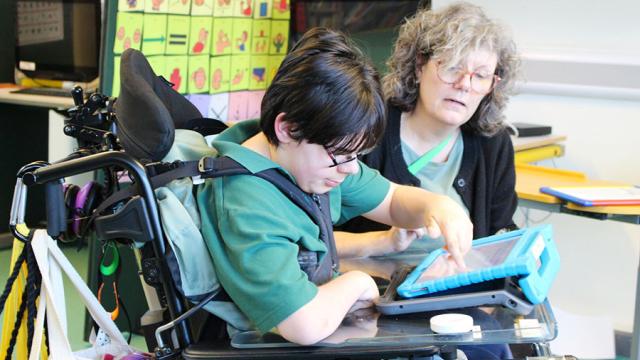Treloar School student using a tablet while his assistant guides him in a classroom setting. the boy is wearing glasses and is very focussed on his task.