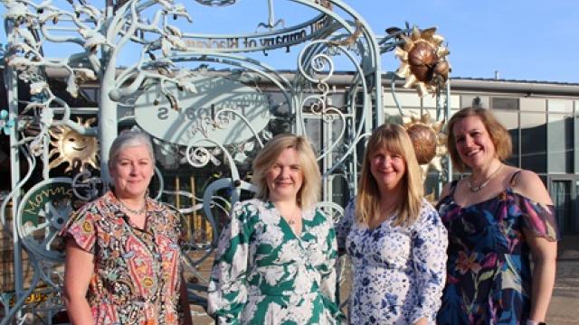 Four women in colorful floral dresses standing outdoors in front of an ornate metal sculpture under a clear blue sky.