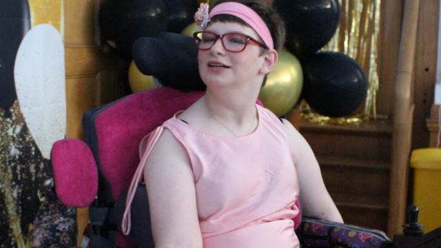 Treloar's student in a pink dress and headband posing for pictures. She is smiling, there are black and gold balloons in the background.