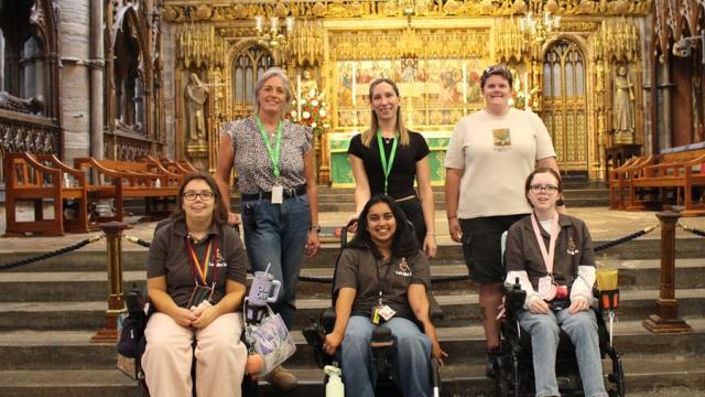 Group of six people, including three in wheelchairs, posing inside a grand cathedral with ornate golden altar in the background.