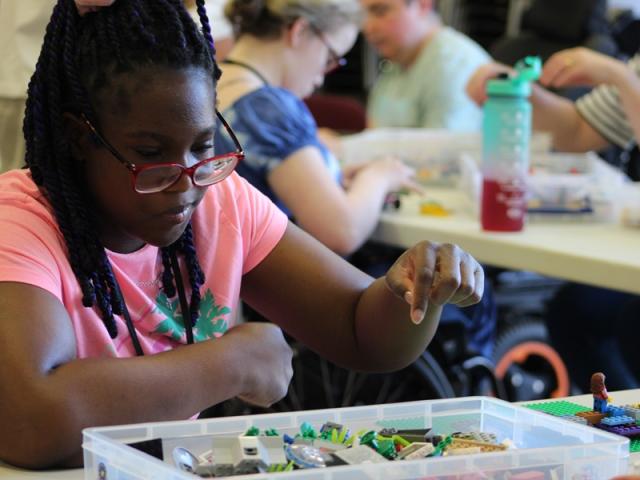 Treloar College student at a table, wearing red glasses; a plastic tray in front of her with Lego pieces. The student is about to pick a piece from the tray.