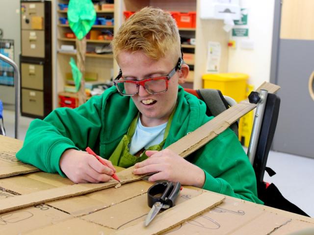 Treloar's student using a pencil to mark where to cut a long piece of cardboard; he is very focused on the task.