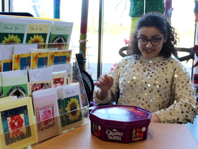 Treloar's student at a stall with student-made cards waiting for customers. She is looking at the camera and smiling.
