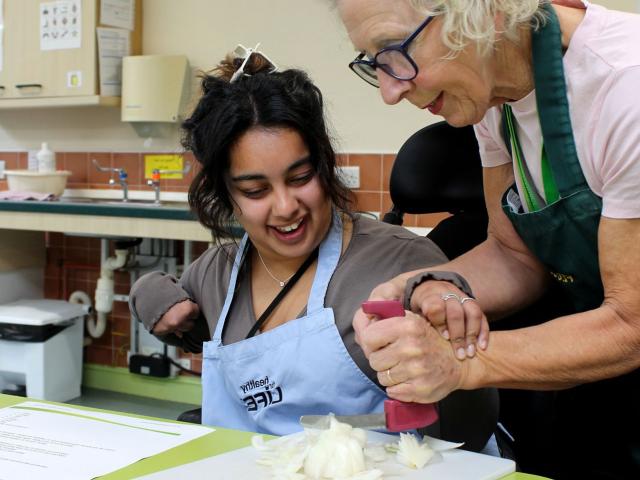 Treloar College student is chopping an onion and her tutor is helping her to do it by holding her hand the the accessible knife.