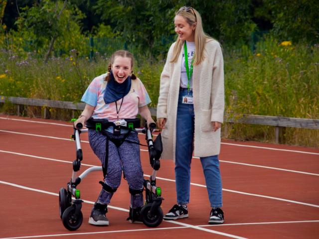 Treloar College student is using a walking on a race track; she is very happy and is smiling; her assistant is standing right next to her, looking at her and smiling.