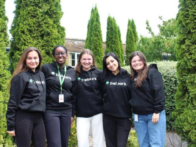 Five female volunteers standing outdoors in front of tall green shrubs, all wearing black hoodies with the 'Treloar's' logo on them.