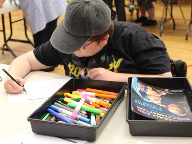 Treloar College student wearing a black cap, drawing with a black marker at a table filled with colorful markers and a book. 