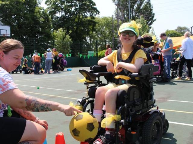 Treloar's primary student wearing yellow accessories plays with a yellow and black soccer ball on an outdoor court during a sunny day during Sports Day. Staff, students and families in the background.