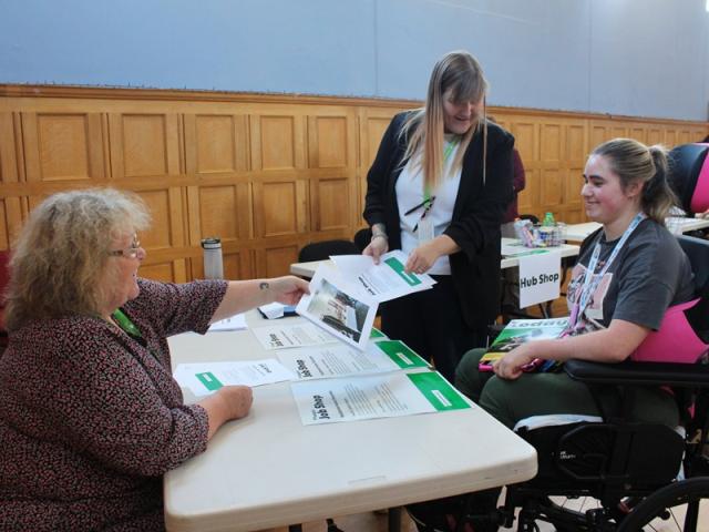 2 Treloar's staff members engage in a discussion with Treloar's student at a table covered with informational brochures inside a wood-paneled room.