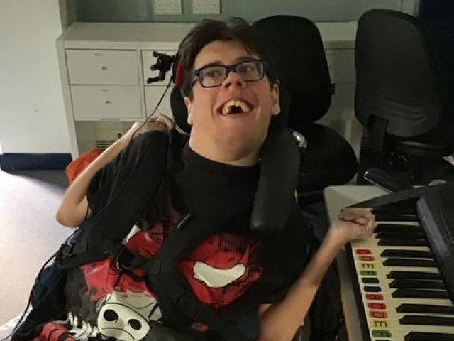 Treloar College student playing the piano with his left hand on the keys; he is wearing glasses and smiling at the camera.
