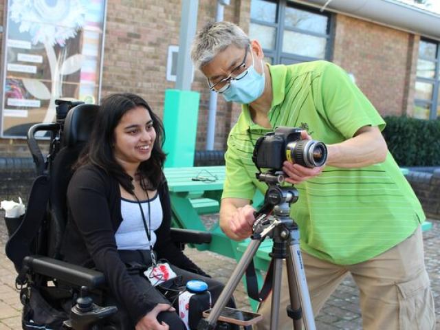 Treloar's student support assistant in green shirt is adjusting a camera on a tripod outdoors for Treloar College student who is about to take some pictures.
