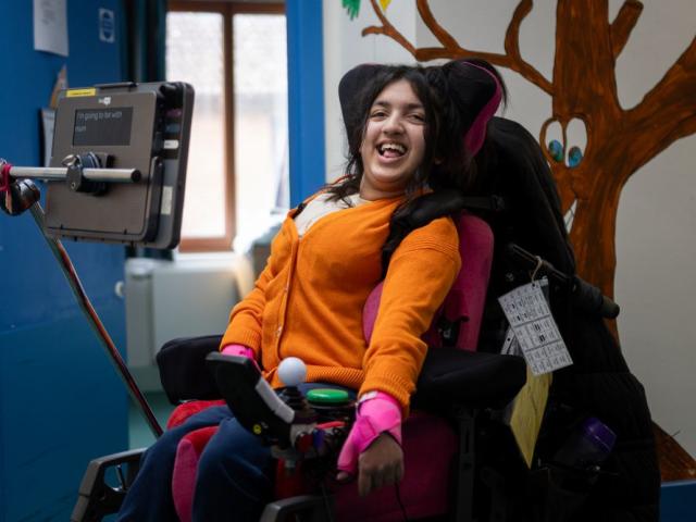 School student in a Powerchair with an AAC Communication device, smiles towards the camera wearing a bright orange cardigan