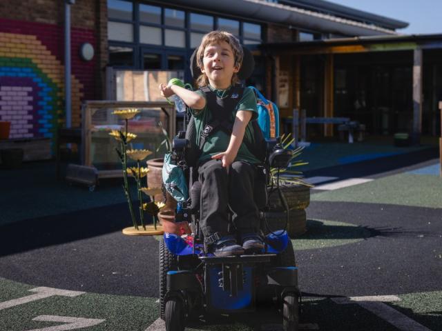 School student in the playground, in the sunshine in a power wheelchair with coloured bricks in a rainbow pattern behind them