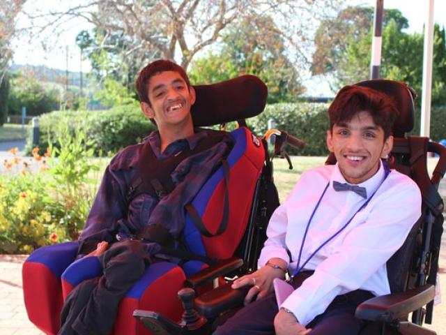 Two male college students sit side by side in their power chairs, smiling and dressed smartly for the College Prom event
