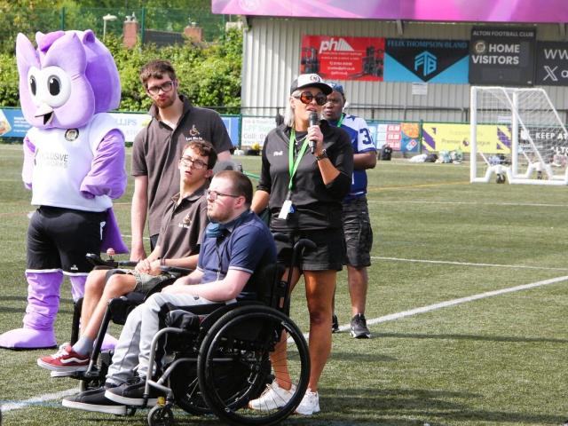Group of people on a football field, including a Treloar's students and staff and a mascot in a purple costume, with one woman speaking into a microphone.