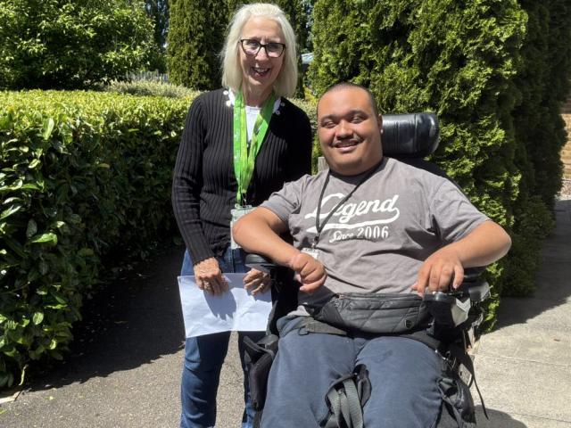 College tutor Flossie stands alongside Student in a wheelchair, both smiling, with trees and hedges behind them and clear blue skies