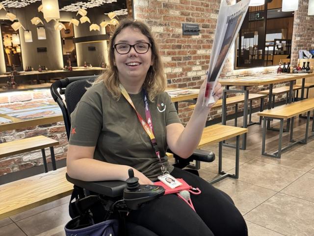 Treloar's student in an empty restaurant holding a big menu and smiling to the camera. She is wearing glasses and a work experience uniform.