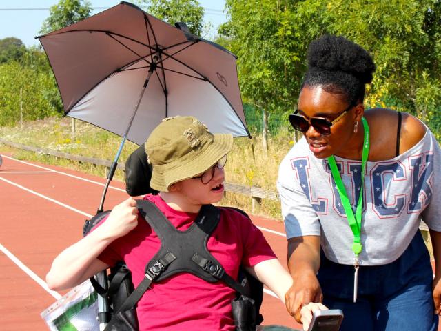 Treloar College student with an umbrella attached to his wheelchair on a sports track chatting with his assistant who is smiling and wearing sunglasses. It is a sunny, warm day.