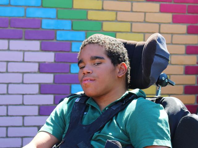 Treloar School student wearing a green T-shirt uniform and poses in front of a colourful brick wall painted in purple, blue, yellow and red hues.