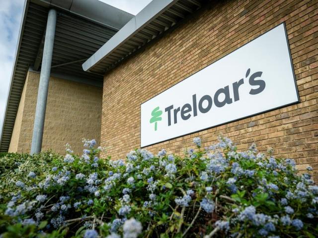 White rectangular sign with 'Treloar's' and a green tree logo mounted on a brick building wall above flowering bushes.