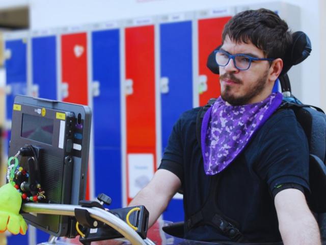 Treloar College student using eye gaze to operate his AAC device with colourful lockers in the background. He is wearing glasses and a black T-shirt.