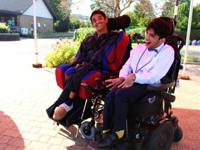 Two Treloar College student smiling and enjoying a sunny day outdoors near a building and garden. They are wearing a formal attire. 