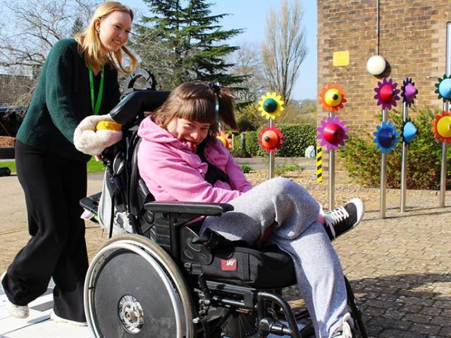 A student support assistant pushes a smiling Treloar College student outdoors near colourful flower-shaped musical instruments on a sunny day.