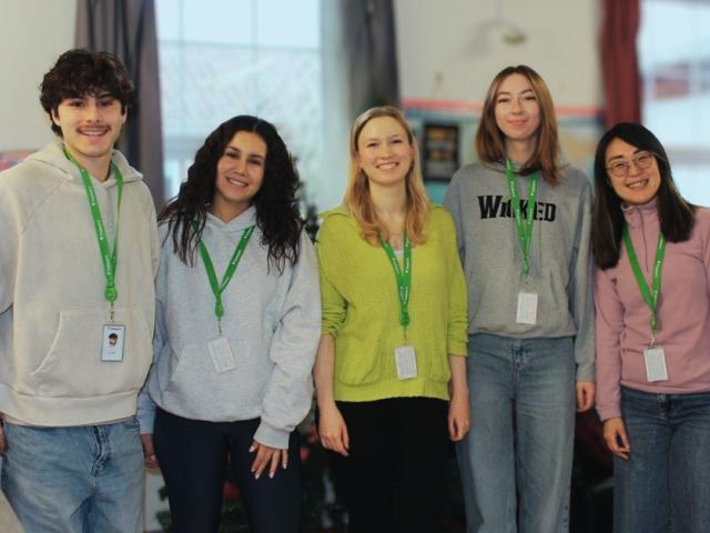 Five young adults wearing ID badges and casual clothes stand smiling in a bright indoor setting.