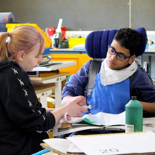 Treloar's student is using a paint brush to paint a stage prop; his assistant is holding his hand to help him paint and hold the brush. they are both really focused on the task.